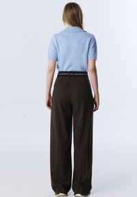 Persoon in een lichtblauw kortärmige gebreide top, ingestopt in een hoogwendige, donkerbruine, wijde broek met een zwarte riem, staande tegen een witte achtergrond.