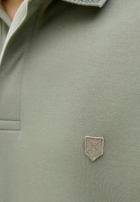 Olivovo zelená polo košeľa s krátkym golierom a textúrovanou béžovou nášivkou na ľavom prsníku. Hladká, elastická tkanina s čistým dizajnom.