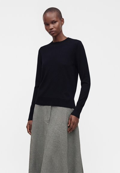 Femme portant un haut noir à manches longues et une jupe grise texturée taille haute, debout devant un fond uni.