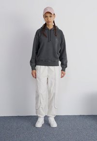 Dark gray hoodie with a kangaroo pocket, paired with white trousers featuring side piping, and white sneakers. Cap is light pink with logo.