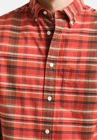 Camicia a quadri rossa con bottoni, con accenti bianchi e blu navy, dotata di taschino sul petto e piccolo ricamo. Realizzata in tessuto di cotone liscio.
