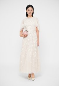 Μακρύ φόρεμα σε κρεμ τούλι με διακοσμητικά λουλούδια και μανίκια με βολάν. Συνδυασμένο με ροζ τσαντάκι και κρεμ παπούτσια. Απαλή υφή.