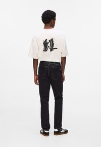 Biely tričko s krátkym rukávom a čiernym grafickým potlačou na chrbte, v kombinácii s čiernymi slim-fit džínsami a čiernymi teniskami s bielymi akcentmi.