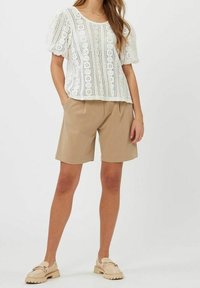 Blusa bianca a occhietti con maniche a sbuffo, abbinata a pantaloni beige plissettati e mocassini chiari. Texture liscia, design casual.