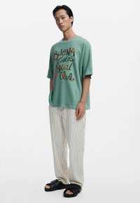 Mint green oversized T-shirt with multicolored text graphic, paired with striped beige pants and black sandals featuring thick straps.