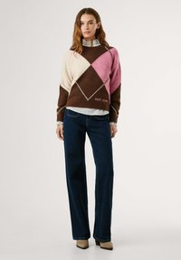 Garen gebreide trui met een roze, bruine en crème ruitpatroon, voorzien van een ribgebreide halslijn en zoom. Gecombineerd met donkerdenim flared jeans.