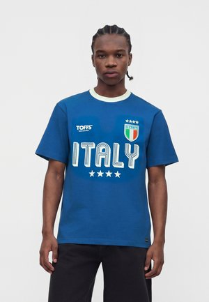 Junger Mann mit geflochtenen Haaren, der ein blaues Italien Fußballtrikot mit Logos und vier Sternen trägt, steht vor einem schlichten hellen Hintergrund.