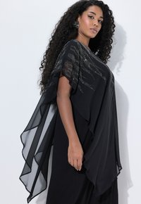 Femme aux longs cheveux bouclés portant une robe noire fluide avec une superposition transparente et ornée, se tenant contre un mur blanc, de profil.