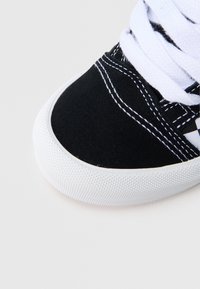 Crna platnena tenisica s bijelim gumenim potplatom, s bijelim vezicama i kontrastnim bijelim šavovima. Zaobljeni vrh s teksturiranim detaljima.