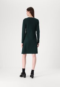 Φόρεμα μακρυμάνικο, σκούρο πράσινο, με ευθεία γραμμή, που φτάνει λίγο πάνω από τα γόνατα. Φορεμένο με μαύρες ψηλοτάκουνες γόβες και κάλτσες μέχρι τον αστράγαλο.