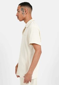 Camisa de manga corta, color beige, con cuello, fabricada en tela acanalada, que presenta un cierre frontal con botones y un diseño liso. Modelo mostrado en vista de perfil.