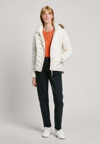 Witte gewatteerde jas met een capuchon, met een ritssluiting, gedragen over een oranje top en zwarte broek, gecombineerd met witte sneakers.