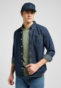Donkerblauw denim overhemd met knopen, een borstzak en opgerolde mouwen, gedragen over een lichtgroen T-shirt. Vergezeld van een marineblauwe baseballcap.
