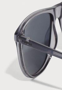 Ochelari de soare gri transparenti cu lentile întunecate, vedere unghiulară care evidențiază designul unghiular al ramei și brandingul "Polaroid" de pe braț.