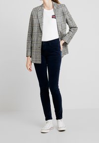 Grå rutiga kavaj med strukturerade axlar, bärs över en vit t-shirt med en liten svart och rosa patch, tillsammans med mörkblå byxor och vita sneakers.