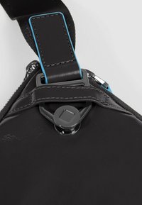 Bolso bandolera negro con acabado liso y brillante. Cuenta con una correa con detalles azules, herrajes metálicos y un cierre seguro con cremallera.