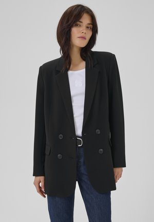 Femeie purtând un sacou negru cu două rânduri de nasturi peste un top alb și blugi albaștri, stând pe un fundal simplu de culoare gri deschis.