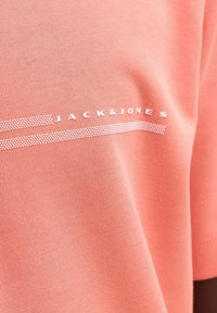 Υφασμάτινο ύφασμα χρώματος κοράλλι με λευκή επιγραφή "JACK & JONES" και δύο διακεκομμένες οριζόντιες γραμμές πάνω και κάτω από το όνομα της μάρκας.