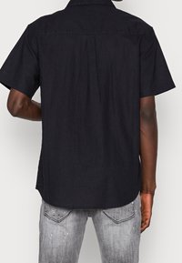 Schwarzes Kurzarmhemd aus Leinen, mit einer Rückenpasse und geradem Saum. Der Stoff hat eine dezente Textur und einen lässigen Schnitt.