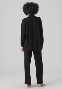 Camicia nera a maniche lunghe con colletto, abbinata a pantaloni larghi neri. Tessuto liscio, con bottoni sulle maniche e vestibilità rilassata.