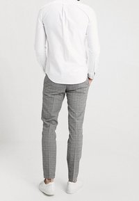 Muž v bílém tričku, šedých kostkovaných slim-fit kalhotách a bílých teniskách, stojící s rukama v kapsách, otočený zády k jednobarevnému pozadí.