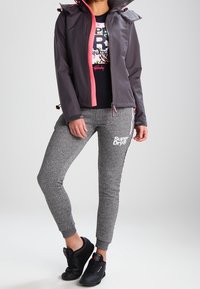 Osoba v černo-růžové bundě, černém tričku s potiskem, šedých teplácích Superdry a černých teniskách, stojící se zkříženýma nohama.