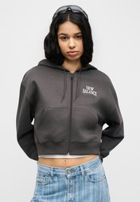 Νεαρή γυναίκα που φοράει σκούρα γκρι ζακέτα με φερμουάρ New Balance, ανοιχτόχρωμο μπλε τζιν και γυαλιά ηλίου τοποθετημένα στο κεφάλι της, ακουμπώντας σε ένα απλό φόντο.