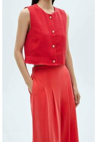 Top corto sin mangas rojo con un acabado texturizado y tres botones metálicos, acompañado de pantalones rojos de cintura alta con piernas anchas.