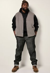 Grau gesteppte Weste über einem schwarzen Hoodie, kombiniert mit dunklen Jeans und braunen Schnürstiefeln. Eine braune Strickmütze rundet den Look ab.