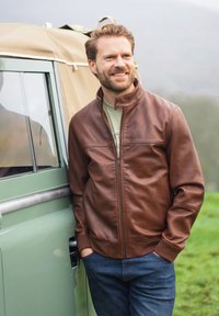 Smiling man with beard wearing brown leather jacket and blue jeans, leaning against a green vintage vehicle outdoors on a foggy day.