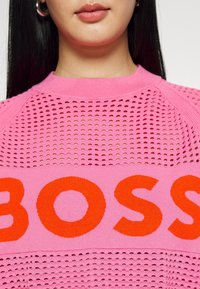 Růžový pletený svetr s průsvitným vzorem a výrazným oranžovým nápisem "BOSS" přes střed. Kulatý výstřih a krátké rukávy.