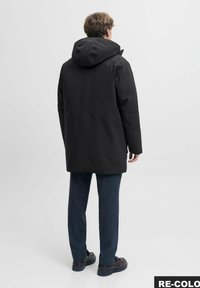 Čierna nepremokavá bunda s kapucňou, ktorá má štíhly dizajn a textúrovanú látku. Nosí sa cez tmavomodré nohavice a čierne topánky.