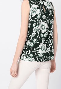 Haut sans manches en tissu vert foncé avec un imprimé floral blanc. Présente un dos à ouverture en clé et une coupe décontractée, associé à un pantalon de couleur claire.