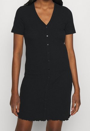 Femme portant une robe noire ajustée à manches courtes avec boutons, col en V et ourlet festonné, debout devant un fond uni.