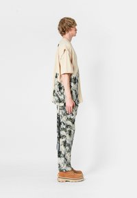 Joven de pie de lado, vistiendo una camisa y pantalones de camuflaje verdes y beige de gran tamaño, con zapatos gruesos color marrón claro, sobre un fondo blanco.