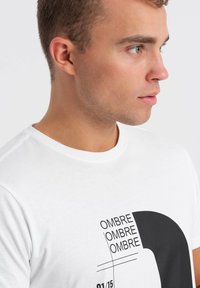 Valkoinen puuvillapaita, jossa on musta graafinen kuvio, jossa lukee "OMBRE" toistuvasti ja pieni numero "01/15" alla. Klassinen pyöreä pääntie.