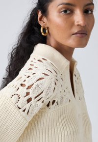 Pull polo en maille crème avec épaules en dentelle tricotée, texture côtelée et col classique.