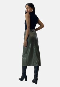 Falda de satén verde oliva con cintura alta, que presenta una abertura en la parte frontal y un diseño sin costuras. Combinada con botas negras de tacón alto hasta la rodilla.