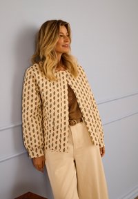 Žena s valovitom plavom kosom koja nosi uzorkovani bež sako, smeđu majicu s čipkom i kremaste hlače širokih nogavica, naslonjena na svijetlosivu zid.