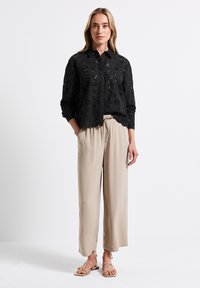 Camicia nera con bottoni, decorata con ricami floreali, maniche lunghe e orlo scallop; abbinata a pantaloni beige larghi e sandali flat.