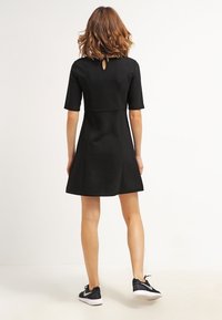 Černé šaty s krátkým rukávem a přiléhavým korzetem, rozevlátou sukní, detailem ve tvaru klíčové dírky na zadní straně, doplněné černými sportovními botami. Hladká textura.