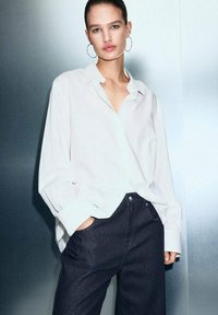Camisa blanca de botones de manga larga con un corte relajado, combinada con unos vaqueros azul oscuro. Textura de tela suave, diseño minimalista y sin patrones visibles.