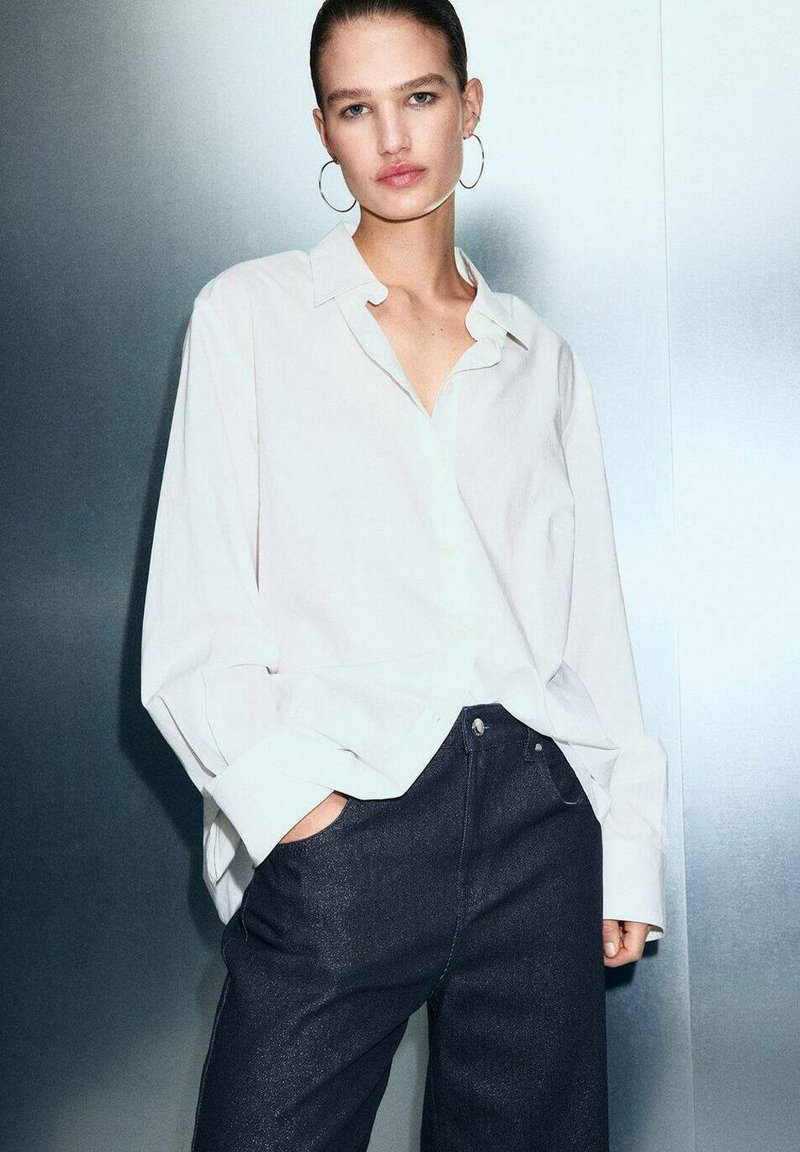 Camisa blanca de botones de manga larga con un corte relajado, combinada con unos vaqueros azul oscuro. Textura de tela suave, diseño minimalista y sin patrones visibles.