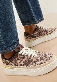 Sneakers na podwyższonej platformie we wzór lamparta, z białymi sznurówkami i złotymi akcentami, noszone z przetarciami niebieskimi dżinsami na jasnej podłodze.