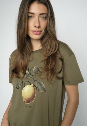 Mladá žena s dlouhými hnědými vlasy, která má na sobě olivově zelené tričko s grafikou citronu a botanickým motivem.