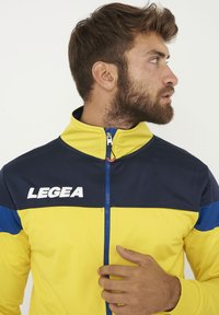 Chaqueta tipo track que presenta una sección superior de color azul marino y una sección inferior de color amarillo brillante, con el logo blanco "LEGEA" y un cierre con cremallera.