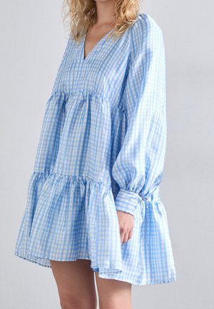 Vrouw draagt een lange mouw, blauw-wit geruiten, vloeiende jurk met een gelaagde structuur en V-hals, staand tegen een effen achtergrond.