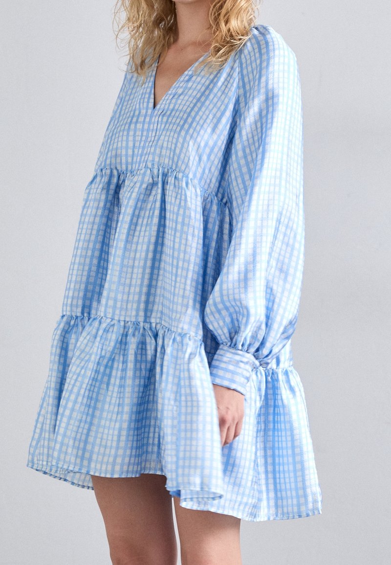 Femme portant une robe fluide à manches longues, à motif à carreaux bleu et blanc, avec un design à volants et un col en V, se tenant devant un fond uni.
