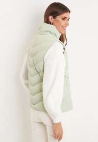 Hellgrüne Pufferweste mit Zickzacknähten, hohem Kragen, kombiniert mit einem weißen strukturierten Pullover, vor einem schlichten Hintergrund präsentiert.