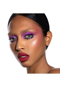 Un primo piano di un volto con ombretto viola vibrante, eyeliner blu e labbra rosse audaci. La pelle ha una texture liscia e un delicato illuminante.